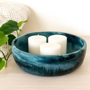 May include: A teal decorative bowl with a marbled effect contains three white pillar candles. A green leafy plant and a cream-coloured folded towel are in the background. The bowl is a decorative item.