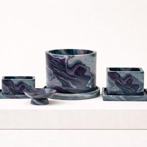 May include: A set of marbled planters and a small dish in shades of blue, purple, and grey. The set includes a large cylindrical planter, two square planters with matching trays, and a small, pedestal dish.