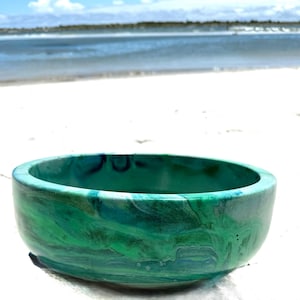 May include: A teal-coloured bowl with a marbled effect, featuring shades of green and blue. The bowl is set on a sandy beach with the ocean in the background. The bowl's design resembles ocean waves.