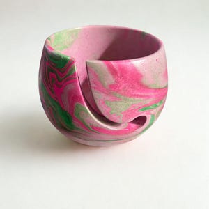 A pink and green marbled yarn bowl. The bowl has a unique design with a curved opening for yarn to feed through. The colors swirl together creating a decorative and functional accessory for knitters and crocheters. The bowl is on a white background. ✨ Handmade Cement Yarn Bowl – swirl design in pink and green, one-of-a-kind finish.