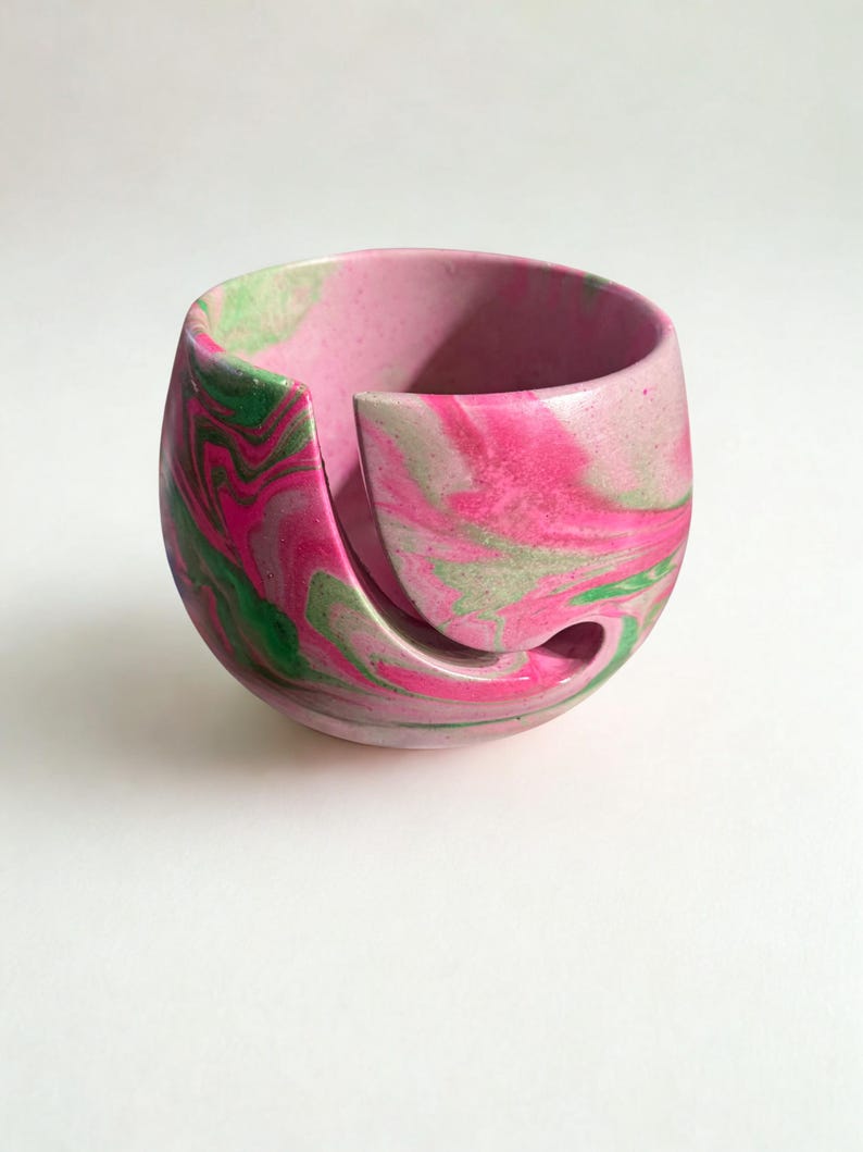 A pink and green marbled yarn bowl. The bowl has a unique design with a curved opening for yarn to feed through. The colors swirl together creating a decorative and functional accessory for knitters and crocheters. The bowl is on a white background. ✨ Handmade Cement Yarn Bowl – swirl design in pink and green, one-of-a-kind finish.