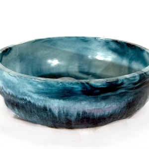 May include: A round, shallow basin with a smooth, glossy interior and a textured exterior. The surface features a blended gradient of deep navy blue at the base, transitioning into lighter shades of teal and turquoise toward the upper rim.