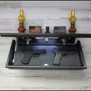 May include: A black wooden wall-mounted gun safe with a hidden compartment. The safe is open, revealing two handguns inside. The safe is decorated with two oil lamps and two American flags.