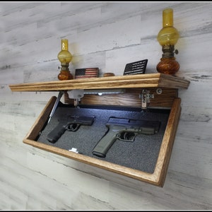 May include: A wooden wall-mounted gun safe with a hidden compartment. The safe is open, revealing two handguns inside. The safe is illuminated by a light inside. The safe is decorated with a small American flag and two oil lamps.