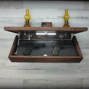 May include: A wooden wall-mounted gun safe with a hidden compartment. The safe is open, revealing two handguns inside. The safe has a light inside and is decorated with a small American flag.