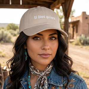 Op de afbeelding: Beige baseballpet met de tekst "Si Dios quiere" in het wit. De persoon draagt een spijkerjasje met bloemenborduurwerk, turkooiskleurige sieraden en een sjaal met patroon.