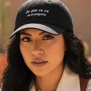 May include: Black baseball cap with the text "Lo que se ve no se pregunta". The cap is worn with a cream-colored button-up shirt, a brown leather vest, and a silver necklace. The background features desert plants.