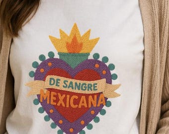 May include: A white t-shirt with a vibrant heart design. The heart includes a flame, a banner with the text "DE SANGRE MEXICANA", and a purple border with green dots. The person is wearing a beige cardigan and blue denim jeans.