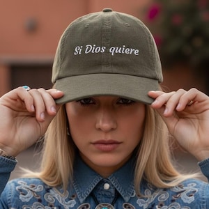 Op de afbeelding: Olijfgroene baseballcap met de tekst "Si Dios quiere" in witte letters. De persoon draagt een spijkeroverhemd met borduursels en een turquoise bolo stropdas.