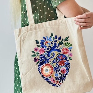 Może przedstawiać: Biała torba tote z płótna z kolorowym wzorem serca z kwiatami i niebieskimi akcentami. Serce jest namalowane w stylu ludowym.