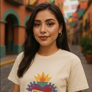 May include: A woman wearing a cream-colored t-shirt with a heart-shaped graphic. The graphic includes a crown, a heart, and the text "DE SANGRE MEXICANA". The background features a colorful street scene with flags.