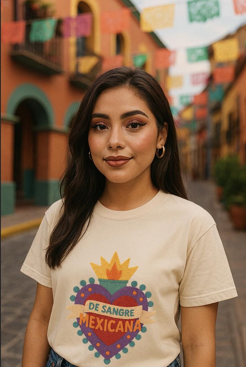 May include: A woman wearing a cream-colored t-shirt with a heart-shaped graphic. The graphic includes a crown, a heart, and the text "DE SANGRE MEXICANA". The background features a colorful street scene with flags.