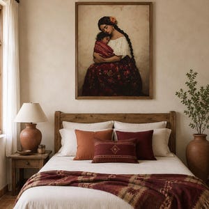 May include: A framed wall art print depicting a person with long dark braided hair holding a child, displayed above a bed with a white duvet, a dark red patterned throw blanket, and several decorative pillows in shades of rust and burgundy.