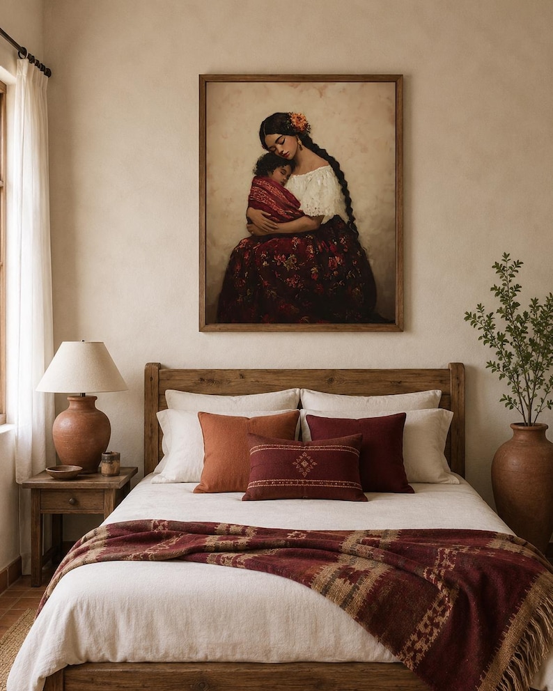 May include: A framed wall art print depicting a person with long dark braided hair holding a child, displayed above a bed with a white duvet, a dark red patterned throw blanket, and several decorative pillows in shades of rust and burgundy.
