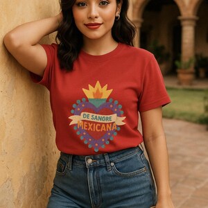 May include: Red t-shirt with a heart-shaped graphic that reads "DE SANGRE MEXICANA". The graphic features a yellow flame and a banner. The model is wearing denim shorts and holding a straw hat.