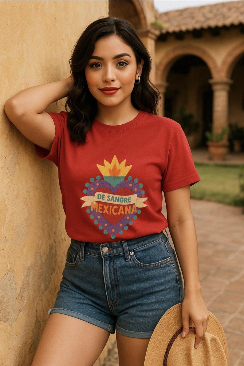 May include: Red t-shirt with a heart-shaped graphic that reads "DE SANGRE MEXICANA". The graphic features a yellow flame and a banner. The model is wearing denim shorts and holding a straw hat.