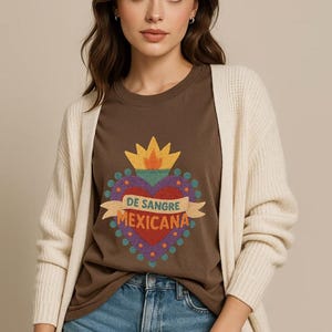May include: A woman wearing a brown t-shirt with a heart graphic and the text "DE SANGRE MEXICANA". She is also wearing a cream-colored cardigan and blue denim shorts. The graphic heart has a crown and is surrounded by colorful dots.