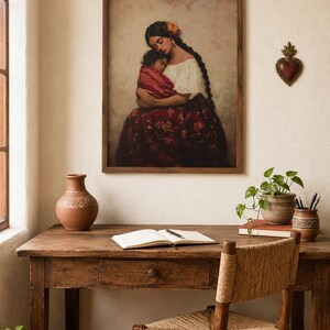 May include: A framed wall art print depicting a person with long dark braided hair holding a sleeping child, both wrapped in a red patterned textile. The person wears a white off-the-shoulder top and a dark red floral skirt against a soft background.
