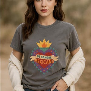 May include: A charcoal gray t-shirt featuring a colorful graphic heart design with the text "DE SANGRE MEXICANA". The design includes a yellow crown, a red heart, and a banner. The model is wearing denim shorts and a cream-colored cardigan.