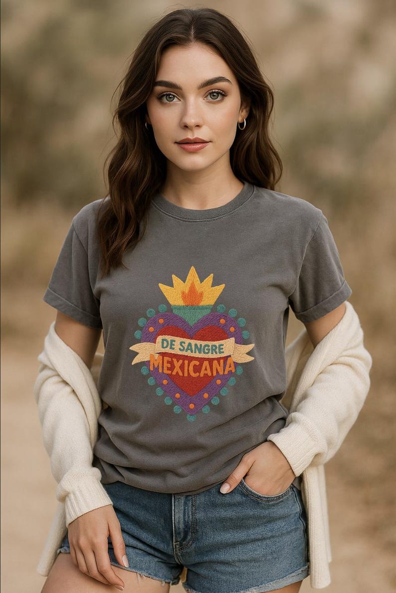 May include: A charcoal gray t-shirt featuring a colorful graphic heart design with the text "DE SANGRE MEXICANA". The design includes a yellow crown, a red heart, and a banner. The model is wearing denim shorts and a cream-colored cardigan.