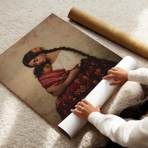 May include: A square art print featuring a painting of a person with long dark braided hair wearing a white top and a dark red floral skirt, holding a sleeping child wrapped in a red patterned cloth against a soft beige background.