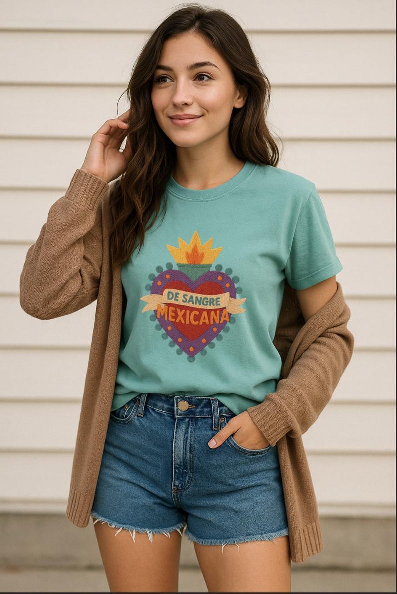 May include: Teal short-sleeve t-shirt with a heart-shaped graphic. The graphic features a red heart, purple border, and a banner with the text "DE SANGRE MEXICANA". The model is wearing denim shorts and a brown cardigan.