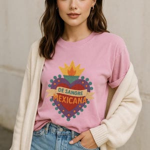 May include: Pink crew neck t-shirt featuring a heart design with the text "DE SANGRE MEXICANA". The heart is red with a yellow flame and is surrounded by a purple border. The shirt is worn with blue jeans and a cream cardigan.