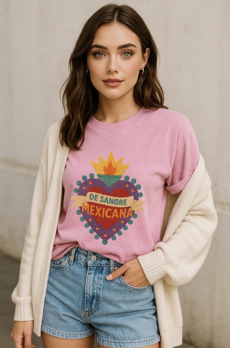 May include: Pink crew neck t-shirt featuring a heart design with the text "DE SANGRE MEXICANA". The heart is red with a yellow flame and is surrounded by a purple border. The shirt is worn with blue jeans and a cream cardigan.
