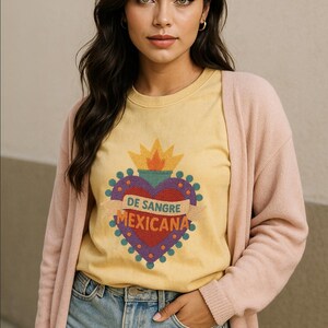 May include: Yellow t-shirt with a heart-shaped graphic that reads "DE SANGRE MEXICANA". The graphic is red, purple, and gold. The person is wearing a pink cardigan and blue denim shorts.