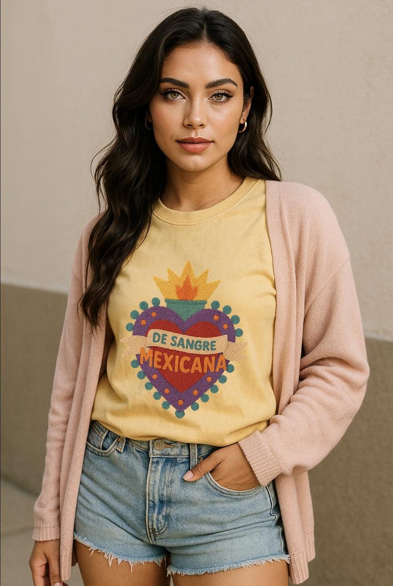 May include: Yellow t-shirt with a heart-shaped graphic that reads "DE SANGRE MEXICANA". The graphic is red, purple, and gold. The person is wearing a pink cardigan and blue denim shorts.