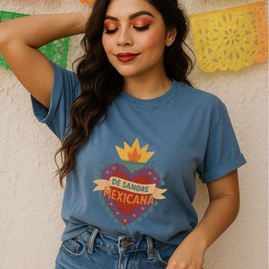 May include: A blue t-shirt with a heart-shaped graphic that reads "DE SANGRE MEXICANA." The graphic is red, purple, and yellow. The person is wearing blue jeans and holding a brown hat. Colorful paper decorations are in the background.