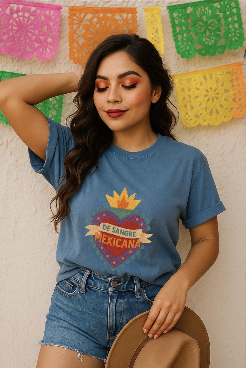 May include: A blue t-shirt with a heart-shaped graphic that reads "DE SANGRE MEXICANA." The graphic is red, purple, and yellow. The person is wearing blue jeans and holding a brown hat. Colorful paper decorations are in the background.