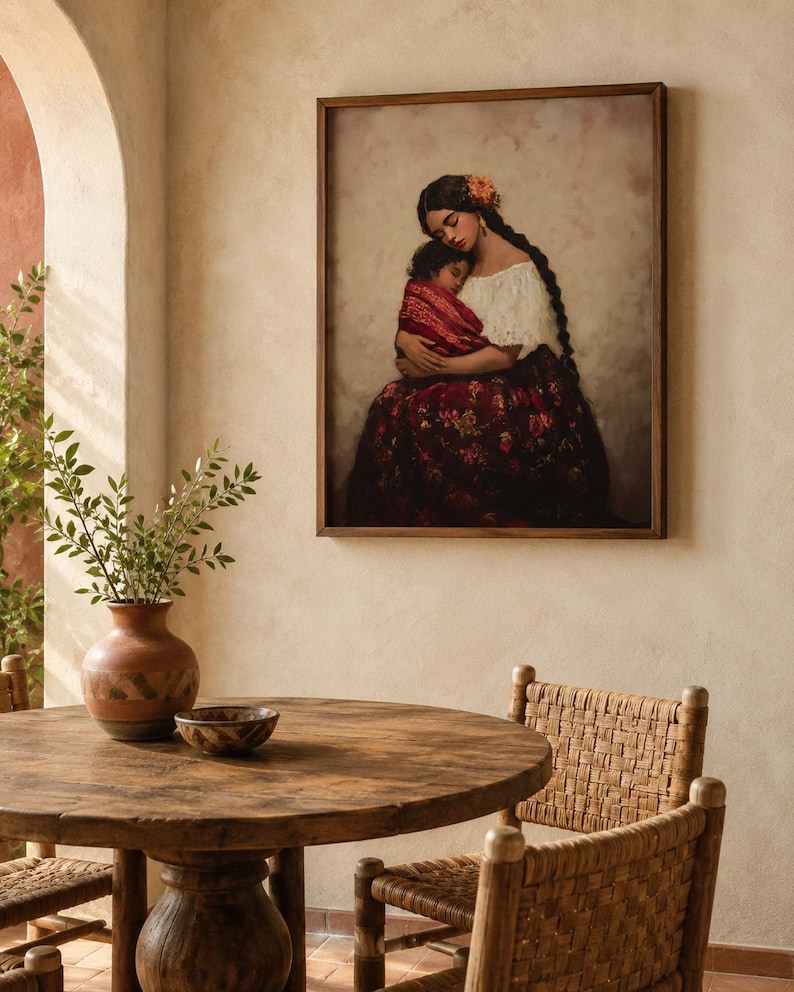 May include: A framed wall art print depicting a person with long dark braided hair wearing a white blouse and a dark red floral skirt, holding a small child wrapped in a red patterned cloth against a soft, neutral-toned background.