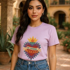 May include: A lilac-colored t-shirt featuring a heart-shaped design with the text "DE SANGRE MEXICANA". The design includes a yellow crown and a banner with the text. The model is wearing blue denim shorts.