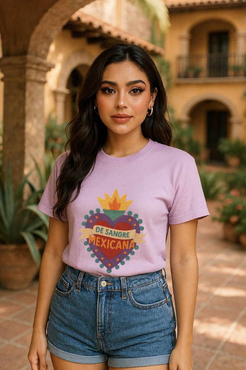 May include: A lilac-colored t-shirt featuring a heart-shaped design with the text "DE SANGRE MEXICANA". The design includes a yellow crown and a banner with the text. The model is wearing blue denim shorts.