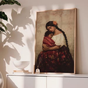 May include: A framed wall art print depicting a person with long dark braided hair and an orange flower accessory, wearing a white off-the-shoulder top and a dark red floral skirt, holding a small child wrapped in a red patterned blanket.