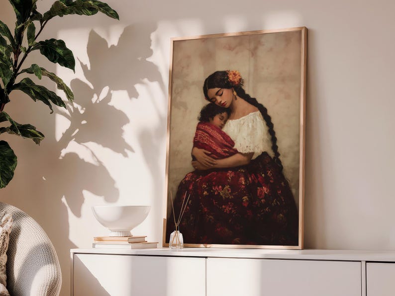 May include: A framed wall art print depicting a person with long dark braided hair and an orange flower accessory, wearing a white off-the-shoulder top and a dark red floral skirt, holding a small child wrapped in a red patterned blanket.