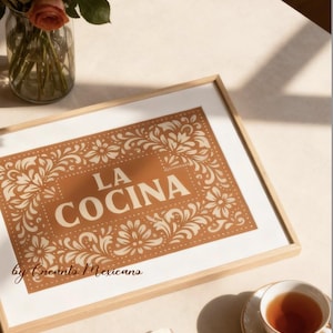 Puede incluir: Impresi&oacute;n art&iacute;stica enmarcada con las palabras "LA COCINA" en una fuente decorativa, rodeada de patrones florales. La impresi&oacute;n est&aacute; en un marco de madera y se encuentra sobre una mesa con una taza de t&eacute;, un libro y un jarr&oacute;n de rosas.