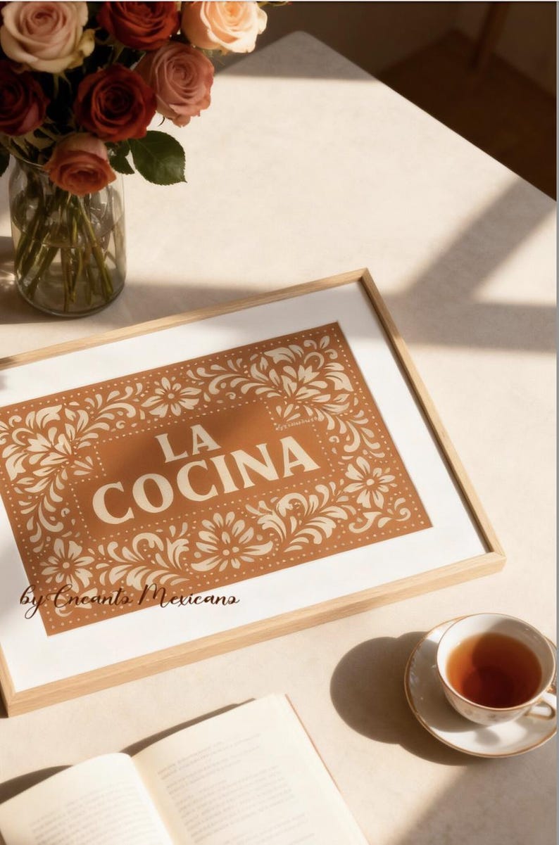 Puede incluir: Impresi&oacute;n art&iacute;stica enmarcada con las palabras "LA COCINA" en una fuente decorativa, rodeada de patrones florales. La impresi&oacute;n est&aacute; en un marco de madera y se encuentra sobre una mesa con una taza de t&eacute;, un libro y un jarr&oacute;n de rosas.
