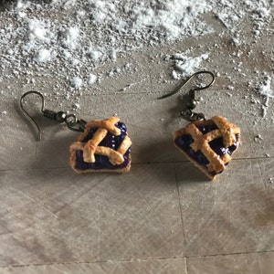 May include: A pair of dangle earrings featuring miniature blueberry pies with a lattice crust. The earrings are made of brown metal and have a hook fastening.