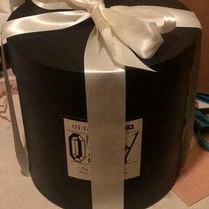 May include: A black cylindrical gift box tied with a wide white satin ribbon and bow. The box has a label with text and a diamond-shaped graphic. The box is on a white surface.