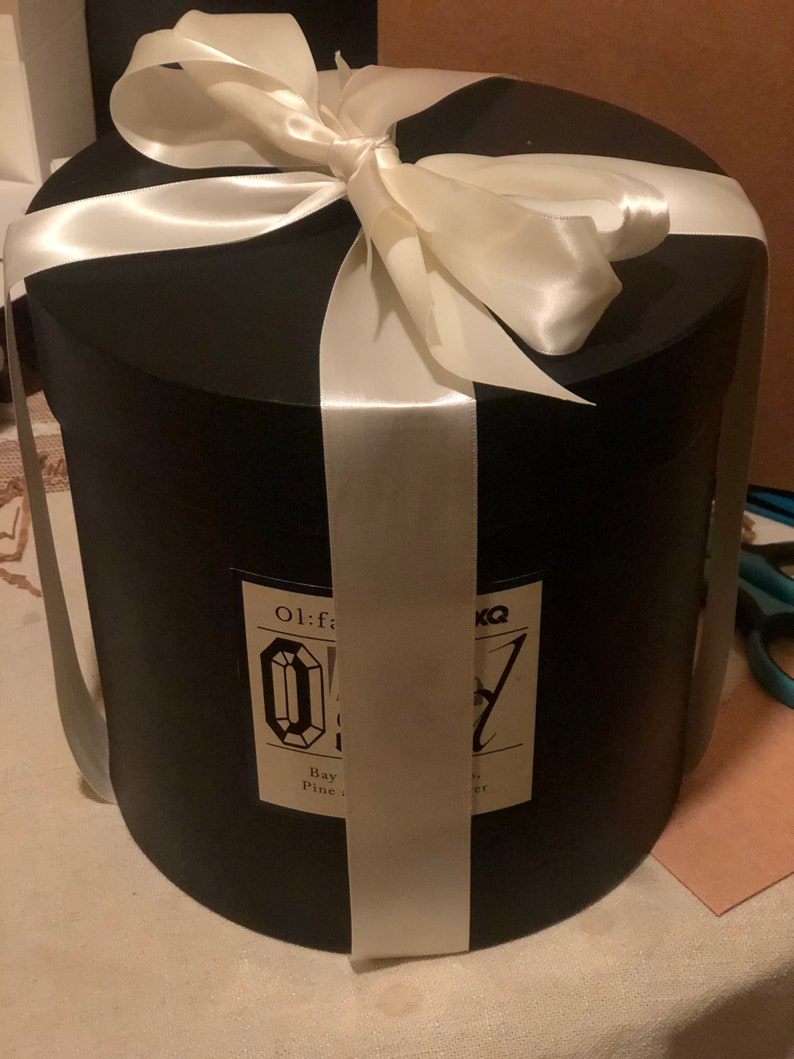 May include: A black cylindrical gift box tied with a wide white satin ribbon and bow. The box has a label with text and a diamond-shaped graphic. The box is on a white surface.