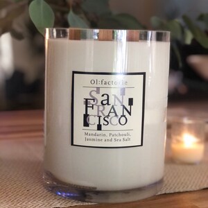 May include: A large, clear glass candle with a silver rim and white wax. The label reads "San Francisco" with scent notes of Mandarin, Patchouli, Jasmine, and Sea Salt. A smaller candle is lit in the background.