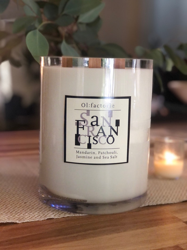 May include: A large, clear glass candle with a silver rim and white wax. The label reads "San Francisco" with scent notes of Mandarin, Patchouli, Jasmine, and Sea Salt. A smaller candle is lit in the background.