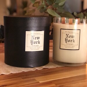 May include: Two scented candles, one in a black cylindrical container and the other in a clear glass. Both have labels that read "New York, The Big Apple, Amberette, Apple and Coriander." The candles are on a wooden surface.