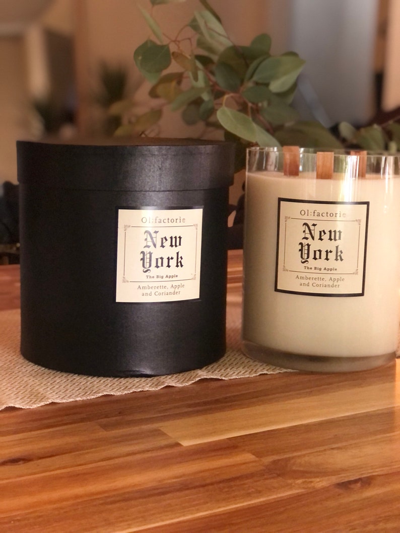May include: Two scented candles, one in a black cylindrical container and the other in a clear glass. Both have labels that read "New York, The Big Apple, Amberette, Apple and Coriander." The candles are on a wooden surface.
