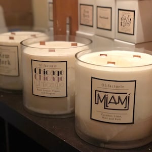 May include: Several large, clear glass candles with wooden wicks are displayed. Each candle has a label with a city name: New York, Chicago, and Miami. The Chicago candle lists basil, fennel, thyme, and oregano as scents. The Miami candle lists coconut, lime, mint, and rum.