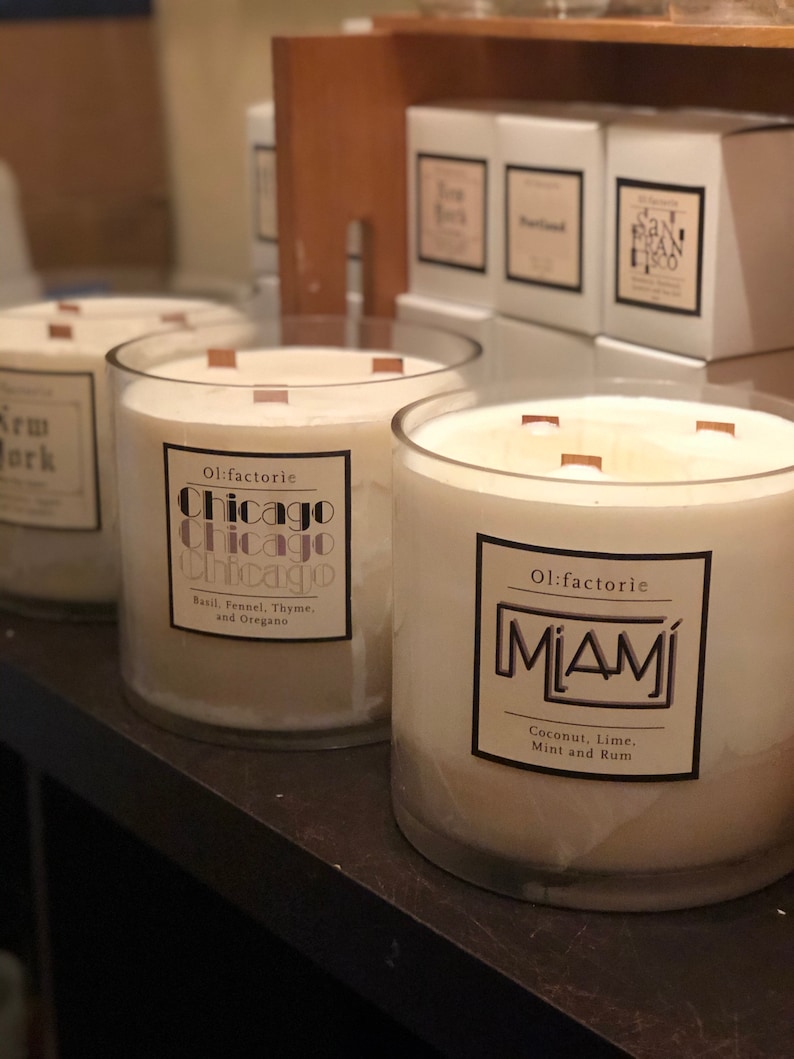 May include: Several large, clear glass candles with wooden wicks are displayed. Each candle has a label with a city name: New York, Chicago, and Miami. The Chicago candle lists basil, fennel, thyme, and oregano as scents. The Miami candle lists coconut, lime, mint, and rum.