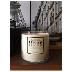 May include: A large, clear glass candle with three wooden wicks. The candle has a white wax and a label that reads "Ol:factorie Chicago" with the scent description: Basil, Fennel, Thyme, and Oregano. The background features a black and white image of a city.