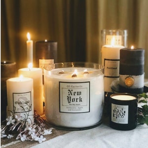 May include: An assortment of lit candles in varying sizes and colors, including white, black, and gray. The candles have labels with city names like "New York," "Oakland," and "San Francisco." Some candles are in glass containers, and the scene is set on a neutral-toned surface.